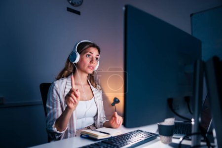 Foto de Mujer de negocios sentada en su escritorio con auriculares con llamada de conferencia mientras trabaja hasta tarde en una oficina - Imagen libre de derechos