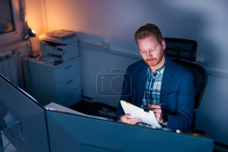 Photo for Confident businessman sitting at his desk reading and signing contracts and financial reports while working late in an office - Royalty Free Image