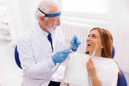 Dentist fixing patient's tooth at dental clinic using dental drill and angled mirror, patient sitting at dental chair and holding saliva ejector for suction