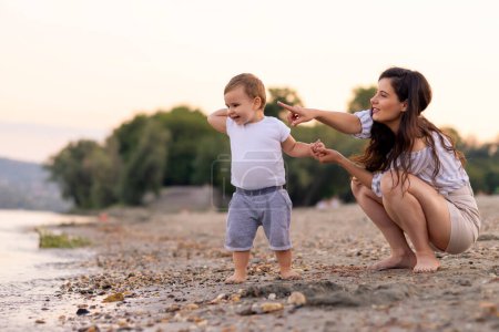 Téléchargez les photos : Belle jeune mère s'amusant à jouer avec son bébé garçon tout en passant l'après-midi d'été sur la plage ensemble, le serrant dans ses bras et jetant des cailloux dans l'eau - en image libre de droit