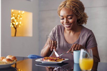 Photo for Beautiful young woman having waffles with fresh berries and chocolate topping for breakfast, enjoying her leisure time at home in the morning - Royalty Free Image