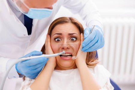 Foto de Mujer asustada mientras está sentada en la silla dental en el consultorio del dentista mientras el médico sostiene el taladro dental y el espejo en ángulo, arreglando el diente del paciente - Imagen libre de derechos