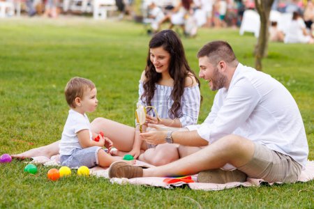 Foto de Lindo niñito sentado en una manta, jugando y comiendo un plátano mientras estaba en un picnic con sus padres en el parque - Imagen libre de derechos