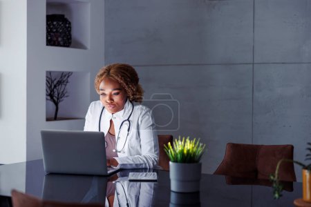 Foto de Doctora sentada en su escritorio en la oficina del hogar, escribiendo informe médico usando computadora portátil mientras tiene consulta de videollamada en línea con el paciente - Imagen libre de derechos