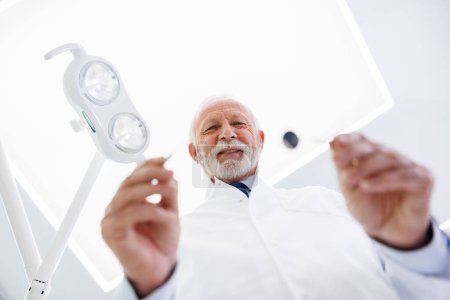 Foto de Vista de ángulo bajo del dentista que sostiene el espejo y la aguja angulados mientras revisa al paciente en la clínica dental - Imagen libre de derechos