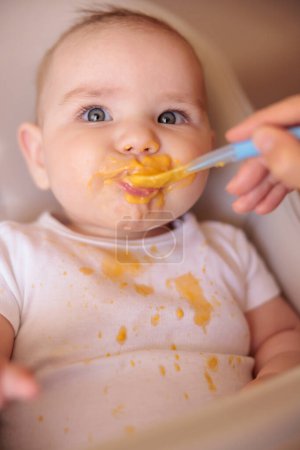 Mère nourrissant la bouillie à son heureux garçon joyeux en utilisant une cuillère, joyeux et heureux bébé assis dans l'appât haut tout désordonné et staied rire
