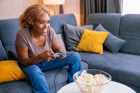 Téléchargez les photos : Belle jeune femme africaine assise sur le canapé du salon s'amusant à passer du temps libre à la maison à jouer à des jeux vidéo - en image libre de droit