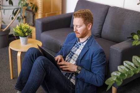 Téléchargez les photos : Développeur logiciel masculin assis sur le plancher du bureau, appuyé sur le canapé et travaillant à l'aide d'un ordinateur portable - en image libre de droit
