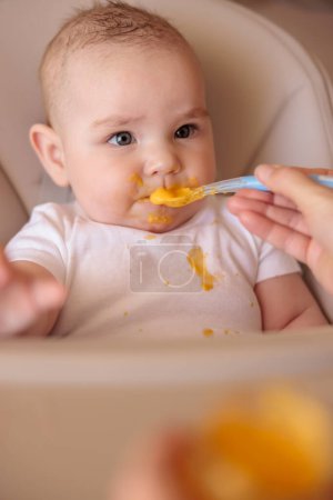 Mère nourrissant le bébé garçon joyeux avec du porridge à la citrouille tout en introduisant de la nourriture solide, bébé assis dans une chaise haute tout sale et staied souriant et mangeant