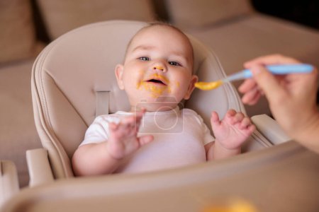 Photo for Mother feeding porridge to her happy cheerful baby boy using spoon, cheerful and happy baby sitting in high chait all messy and staied laughing - Royalty Free Image
