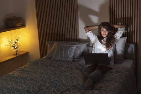 Foto de Mujer sentada en la cama tarde en la noche, sosteniendo el ordenador portátil en su regazo, divirtiéndose mientras ve la película en línea - Imagen libre de derechos