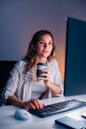 Photo for Female web designer working late in the office, drinking coffee and using laptop computer; casual business woman working overtime - Royalty Free Image