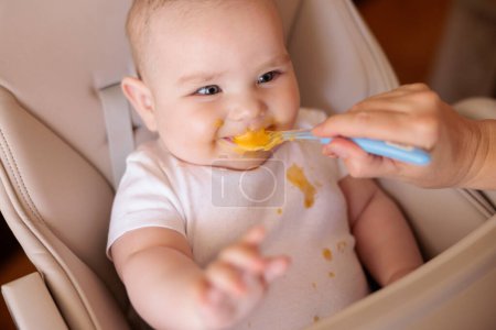 High angle view of mother feeding cheerful baby boy with porridge, baby sitting in high chair all messy and staied smiling and eating