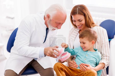 Photo for Senior male dentist educating child patient sitting in mother's lap about proper teeth brushing techniques using plastic human jaw model and tooth brushes - Royalty Free Image