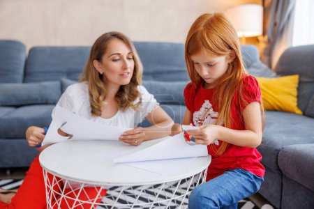 Photo for Beautiful mother helping her little daughter with school project for homework, folding and cutting paper using scissors - Royalty Free Image
