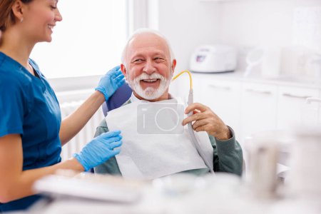 Photo for Senior man sitting at dentist chair holding saliva ejector while doctor fixing his tooth - Royalty Free Image
