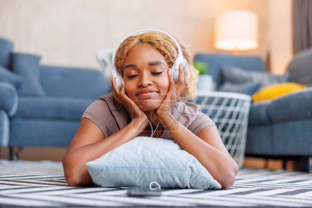 Téléchargez les photos : Belle femme couchée sur le sol, portant un casque, souriant et écoutant la musique tout en se relaxant à la maison - en image libre de droit
