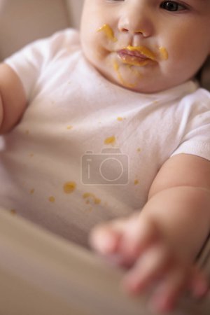 Adorable little baby boy sitting in high chair all messy and stained after eating porridge