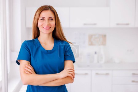 Photo for Portrait of young female doctor wearing uniform standing in ER and smiling with hands crossed - Royalty Free Image