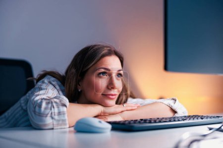 Foto de Mujer de negocios casual sentada en su escritorio en una oficina, trabajando horas extras tarde en la noche usando computadora de escritorio; programadora femenina trabajando horas extras - Imagen libre de derechos