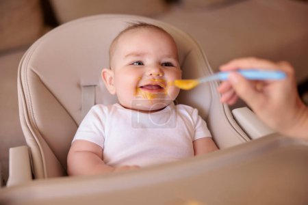 Mère nourrissant son joyeux bébé garçon à l'aide d'une cuillère, introduisant le premier repas solide, bébé assis dans une chaise haute tout désordonné et empilé de bouillie, souriant et mangeant