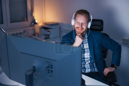 Téléchargez les photos : Homme d'affaires assis à son bureau portant casque ayant réunion en ligne tout en travaillant tard dans un bureau, souriant - en image libre de droit