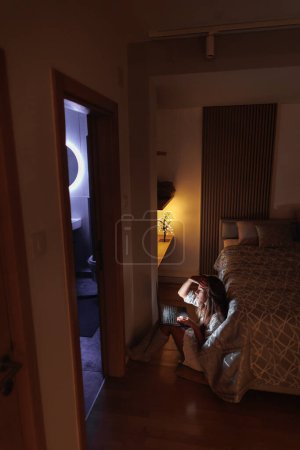 Foto de Freelancer sentada en el suelo junto a la cama usando un ordenador portátil mientras trabaja hasta tarde por la noche - Imagen libre de derechos