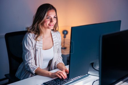 Photo for Casual businesswoman sitting at her desk in an office, working overtime late at night using desktop computer; female programmer working overtime - Royalty Free Image