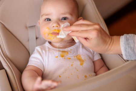 Mother wiping baby using wet wipes after a meal, introducing first solid food meal, baby sitting in high chair all messy and staied with porridge, smiling