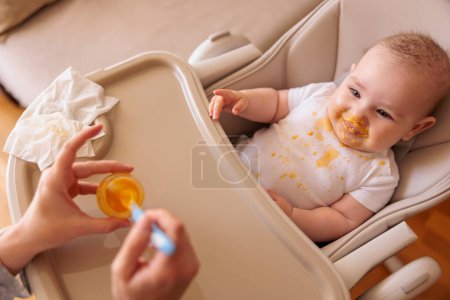High angle view of mother feeding cheerful baby boy with porridge, baby sitting in high chair all messy and staied smiling and eating