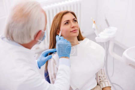 Mujer que tiene tratamiento de belleza anti-edad en el consultorio del médico; médico que aplica rellenos dérmicos a la cara del paciente femenino, corrigiendo arrugas