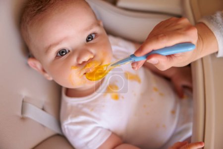 High angle view of mother feeding porridge to baby boy using spoon, introducing first solid food meal, angry baby sitting in high chair refusing food