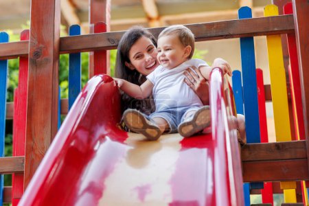 Photo for Beautiful young mother and cute baby boy having fun playing on a slide in the park, mother helping toddler son to climb on the top - Royalty Free Image