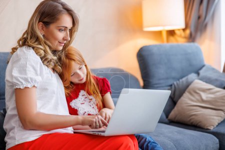 Mother tutoring daughter at home and helping her with homework using laptop computer for online lessons; homeschooling and distance learning concept