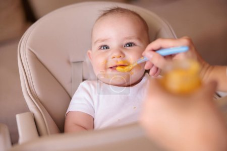Téléchargez les photos : Mère nourrissant la bouillie à son heureux garçon joyeux en utilisant une cuillère, joyeux et heureux bébé assis dans l'appât haut tout désordonné et staied rire - en image libre de droit