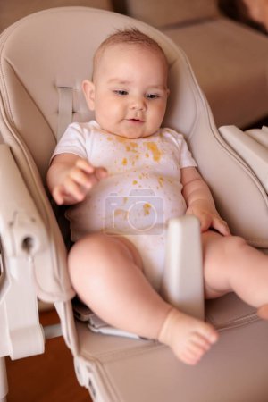 Foto de Lindo bebé niño riendo y jugando en la silla alta todo desordenado y manchado después de comer gachas - Imagen libre de derechos