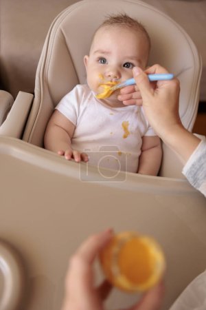 Vue grand angle de la mère nourrissant mignon petit garçon avec de la bouillie, bébé assis dans la chaise haute tous salissant et staied souriant et mangeant