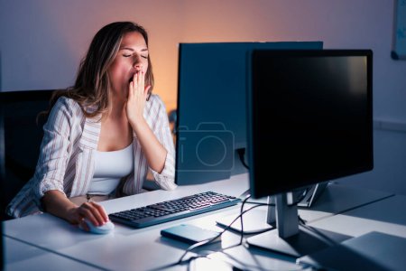 Photo for Woman yawning while sitting at her office desk, tired while working overtime late at night - Royalty Free Image