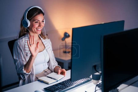 Photo for Business woman sitting at her desk wearing headset having conference call while working late in an office - Royalty Free Image