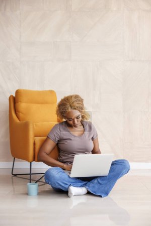 Foto de Freelancer sentada en el suelo trabajando desde casa usando computadora portátil y bebiendo café; mujer trabajando remotamente desde casa - Imagen libre de derechos
