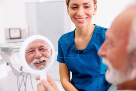 Téléchargez les photos : Homme âgé regardant le miroir et souriant après le check-up au cabinet du dentiste ; dentiste et patient à la clinique dentaire - en image libre de droit