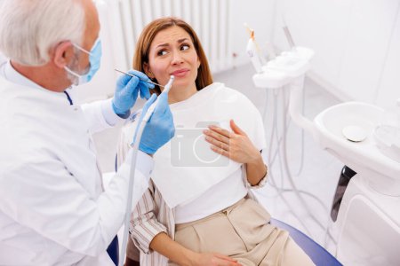 Photo for Woman afraid while sitting at dental chair at dentist office while doctor is holding dental drill and angled mirror, fixing patient's tooth - Royalty Free Image