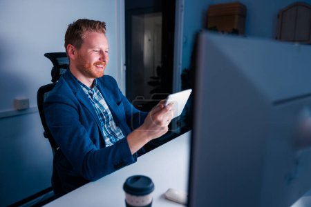 Téléchargez les photos : Homme d'affaires occasionnel utilisant un ordinateur tablette tout en faisant des heures supplémentaires dans un bureau tard dans la nuit - en image libre de droit