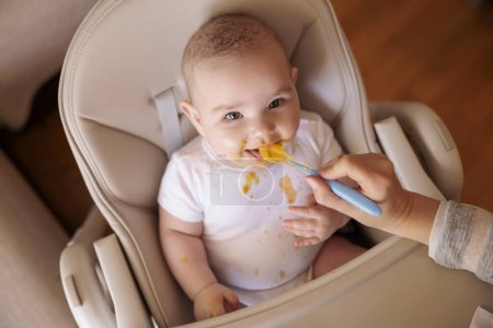 Foto de Vista de ángulo alto de la madre alimentando al niño alegre con gachas usando cuchara, el bebé sentado en la silla alta todo desordenado y estacionado sonriendo y comiendo - Imagen libre de derechos