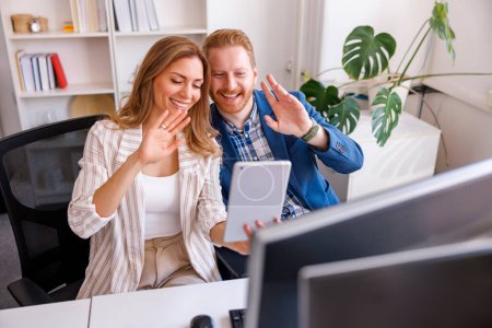 Foto de Gente de negocios teniendo una reunión en línea usando tableta mientras trabajan juntos en una oficina, sonriendo y saludando a la cámara - Imagen libre de derechos