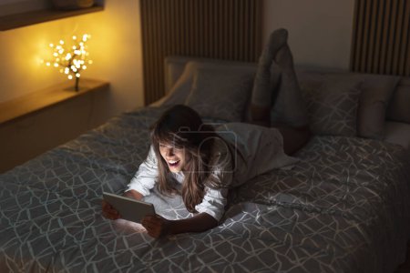 Foto de Hermosa mujer acostada en la cama tarde en la noche, teniendo videollamada usando tableta - Imagen libre de derechos