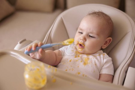 Cheerful funny baby boy sitting in high chair, holding spoon and playing all stained while eating pumpkin porridge from a jar