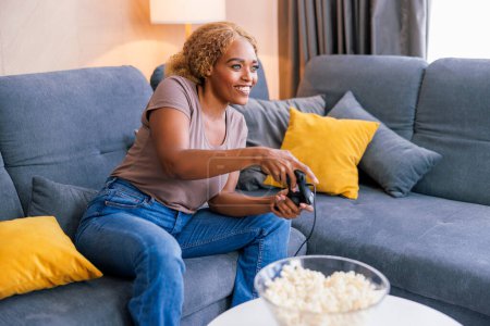 Téléchargez les photos : Belle jeune femme africaine assise sur le canapé du salon s'amusant à passer du temps libre à se détendre à la maison en jouant à des jeux vidéo - en image libre de droit