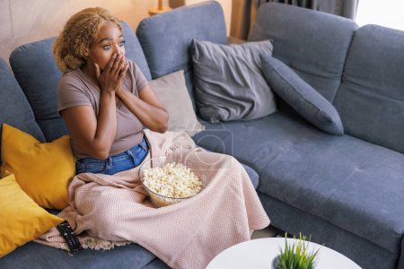 Photo for High angle view of beautiful young african woman sitting on living room couch, eating popcorn and watching horror movie on TV, shocked and scared - Royalty Free Image