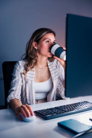 Foto de Diseñadora web femenina trabajando hasta tarde en la oficina, bebiendo café y usando computadora portátil; mujer de negocios ocasional trabajando horas extras - Imagen libre de derechos